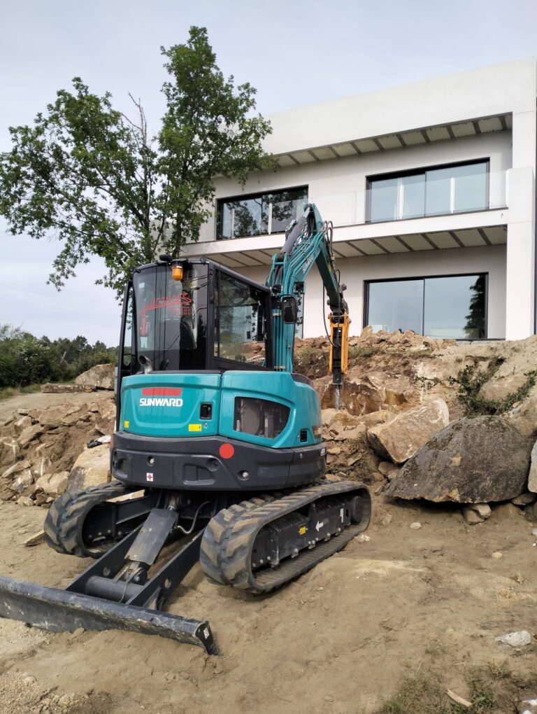 Excavadora de Alucondes con martillo hidráulico trabajando junto a una vivienda